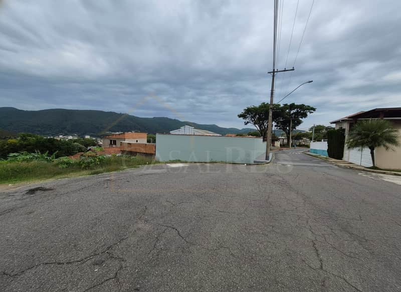 Rua asfaltada com casas e montanhas ao fundo. Céu nublado. Área externa residencial.