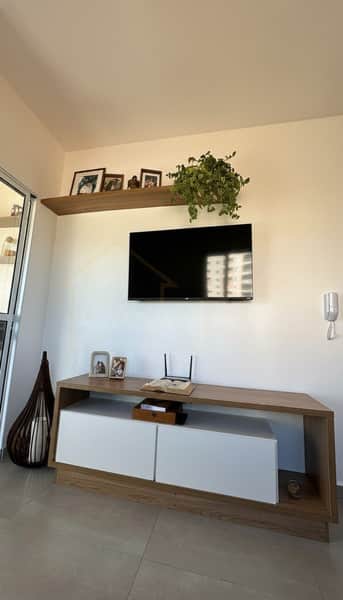 Sala decorada com TV, móvel amadeirado, prateleira com quadros e planta pendente. Ambiente clean e moderno.