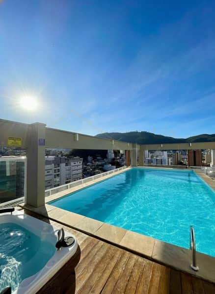 Área externa luxuosa com piscina azul e hidromassagem em deck de madeira, vista panorâmica da cidade e montanhas.