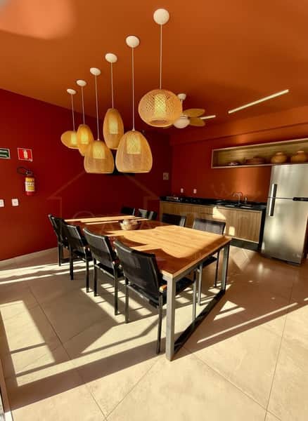Sala de jantar moderna com mesa de madeira, cadeiras pretas e luminárias pendentes de palha. Cozinha ao fundo.