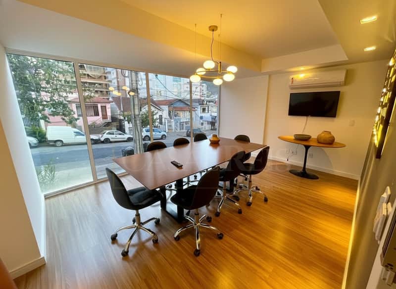 Sala de reunião moderna com mesa, cadeiras pretas, piso de madeira e iluminação elegante. Janelas amplas com vista para a rua.