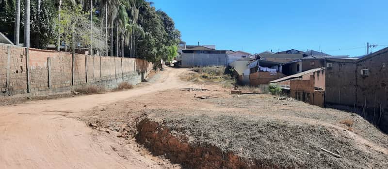 Terreno baldio com casas ao fundo e muro de tijolos. Área externa ensolarada com vegetação seca.