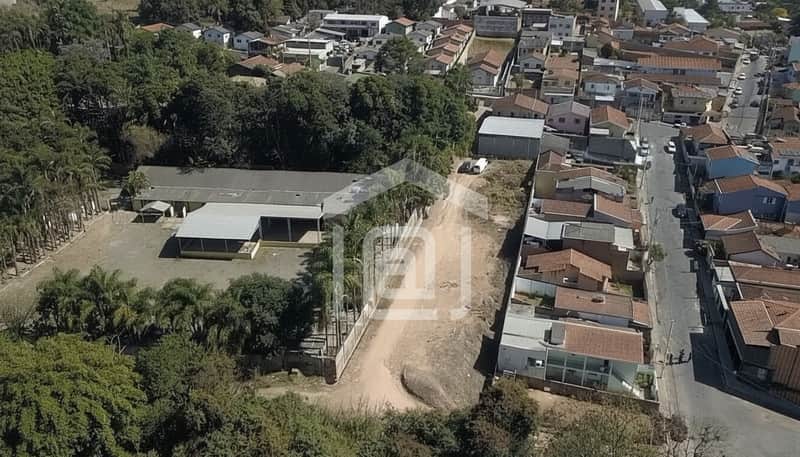 Visão aérea de terreno urbano com galpão, vegetação e casas ao fundo. Espaço amplo e ensolarado.