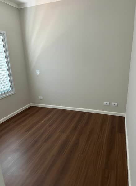 Quarto vazio com piso amadeirado e paredes claras. Janela com persiana à esquerda. Ambiente iluminado e clean.