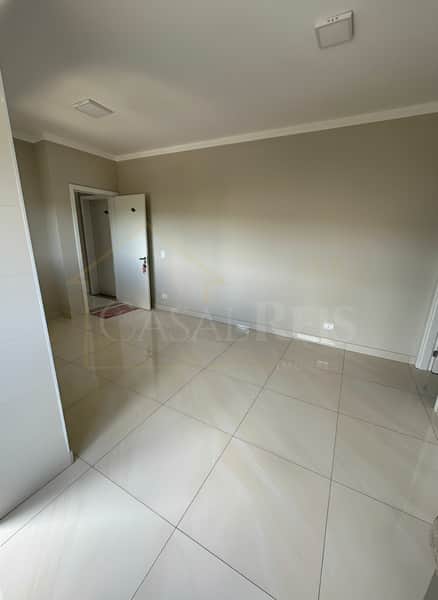 Quarto amplo e claro com piso cerâmico, paredes neutras e porta para outro cômodo. Iluminação embutida no teto.