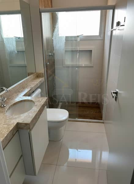 Banheiro claro com pia em mármore, vaso sanitário branco e box de vidro com nicho. Piso brilhante e porta branca.