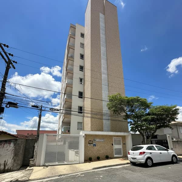 Fachada de prédio residencial alto com varandas e portão branco. Carro estacionado na rua. Céu azul.