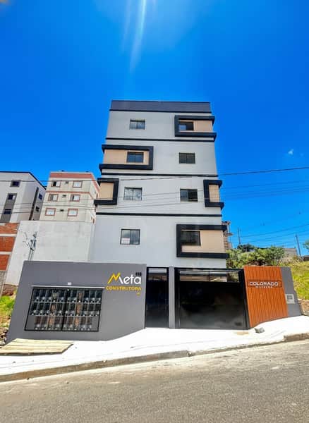 Fachada de prédio moderno com janelas geométricas e portão preto. Destaque para o céu azul e a luz solar.