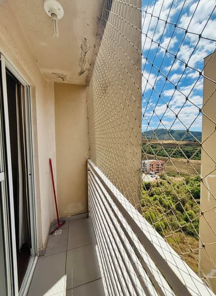Varanda com piso cinza, rede de proteção e vista para área verde. Paredes claras e iluminação no teto.