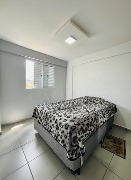 Quarto com cama de casal e manta estampada. Piso cerâmico e janela com veneziana. Iluminação central.