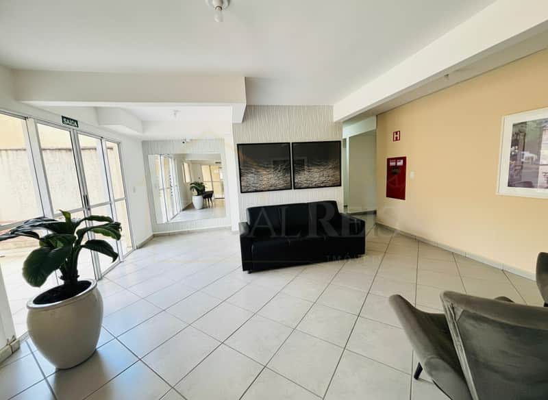 Sala de espera com sofá preto, quadros, planta e piso cerâmico claro. Ambiente iluminado e clean.