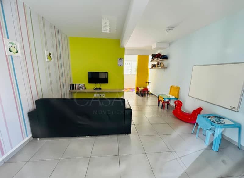 Sala infantil colorida com sofá preto, TV, quadro branco e brinquedos. Ambiente alegre e convidativo.