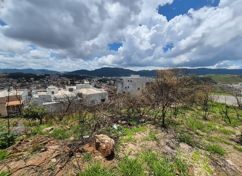 Terreno com vegetação rasteira e vista para a cidade. Construções ao fundo e céu com nuvens.