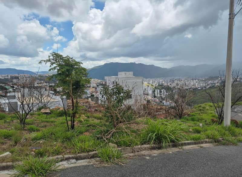 Terreno com vista para cidade e montanhas. Vegetação rasteira e árvores. Céu nublado.