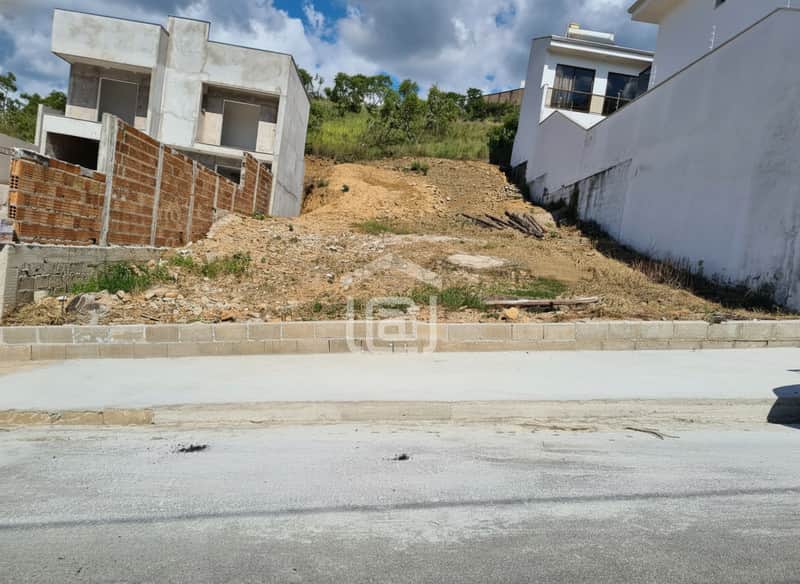 Terreno em declive com construção inacabada. Vegetação rasteira e casas ao fundo. Rua asfaltada.