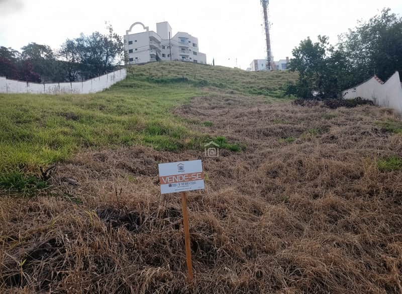 Terreno à venda em declive com vegetação rasteira. Placa "Vende-se" em primeiro plano. Edifício ao fundo.
