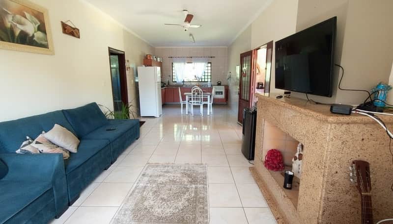 Sala ampla com sofá azul, TV, lareira rústica e piso cerâmico claro. Cozinha ao fundo.