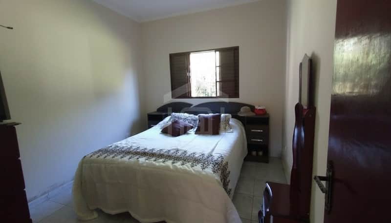Quarto aconchegante com cama de casal, cabeceira escura, janela e piso cerâmico claro. Decoração simples e iluminação natural.