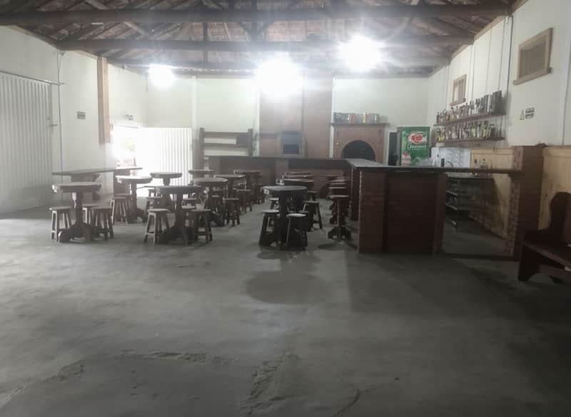 Salão amplo com mesas de madeira rústica, balcão de tijolos e churrasqueira ao fundo. Ambiente iluminado.