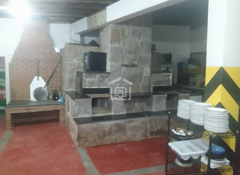 Cozinha rústica com forno de pedra, bancada de tijolos e piso vermelho. Prateleiras com louças. Decoração aconchegante.
