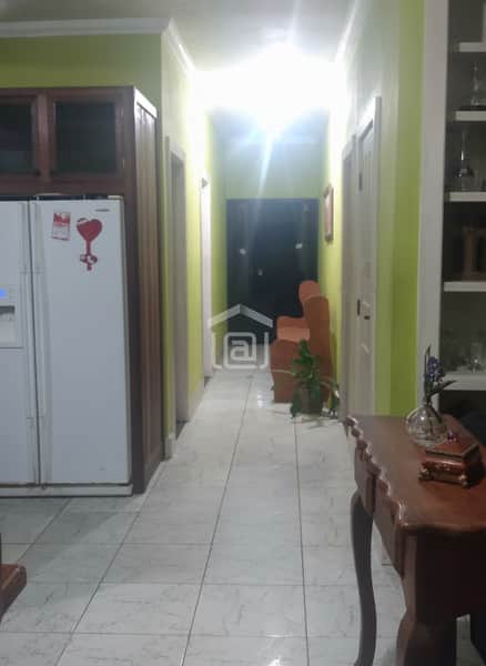 Interior de imóvel com piso cerâmico, geladeira branca e corredor iluminado com porta escura ao fundo.