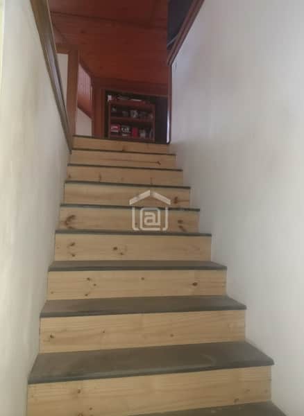 Escada de madeira clara com detalhes escuros. Ambiente interno com paredes brancas e teto amadeirado.