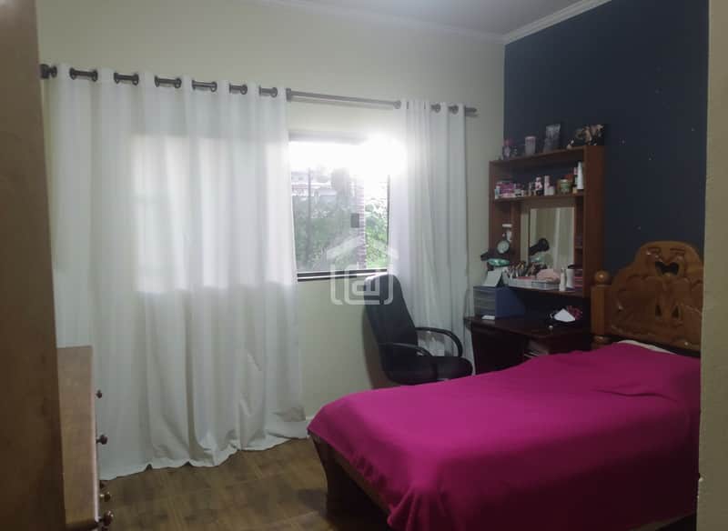Quarto aconchegante com cama rosa, cortina branca na janela e escrivaninha de madeira escura.