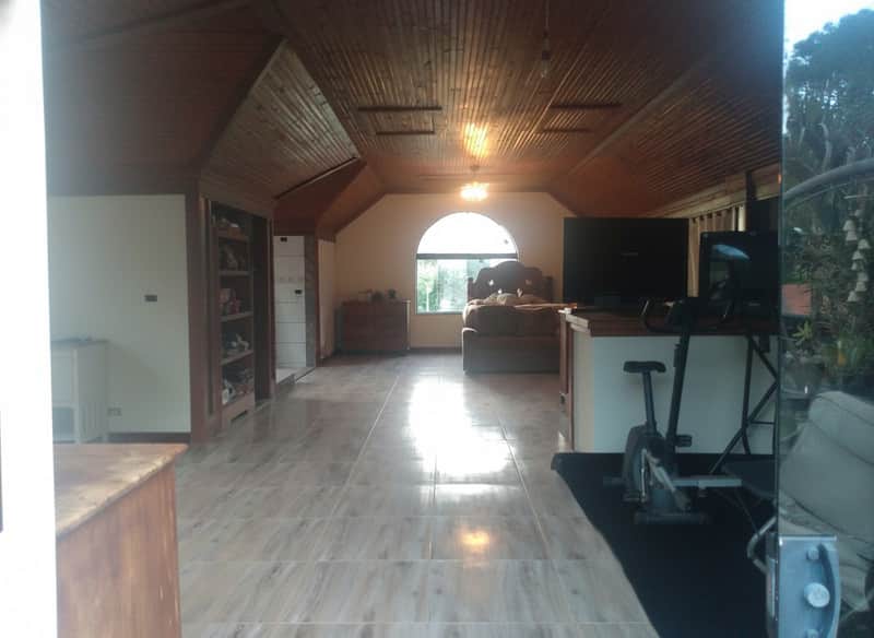 Quarto amplo com cama, TV, bicicleta ergométrica e teto de madeira. Iluminação natural e piso cerâmico.