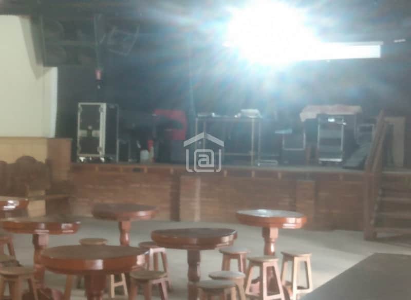 Salão amplo com palco, mesas e cadeiras de madeira. Iluminação forte e equipamentos de som visíveis.