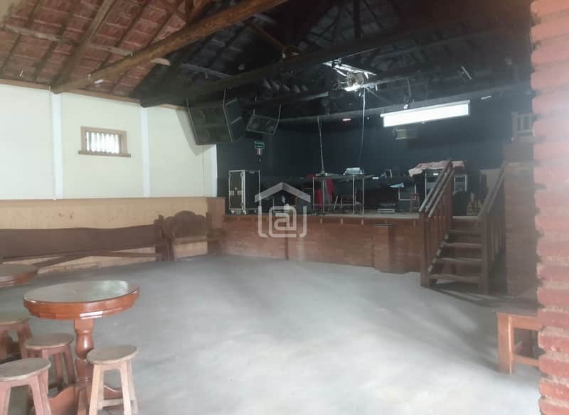 Salão amplo com palco, mesas e cadeiras rústicas. Ambiente iluminado e espaçoso.