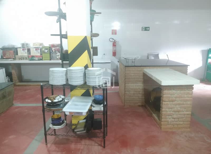 Cozinha industrial ampla com ilha em tijolinho, prateleiras com louças e piso vermelho. Ambiente funcional.