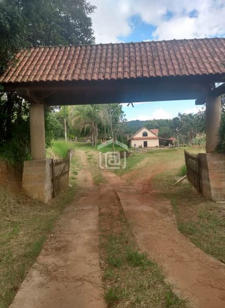 Entrada de sítio com portão rústico de madeira e telhas. Casa bege ao fundo, gramado e árvores.