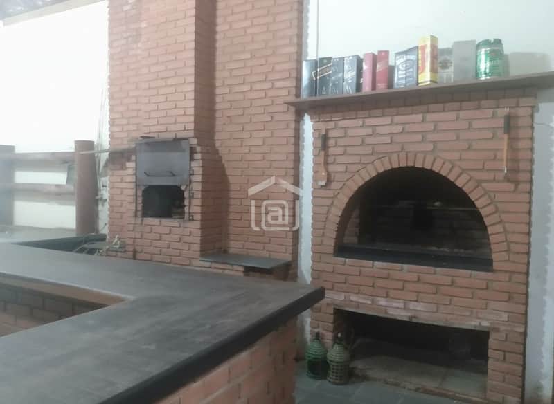 Área externa com churrasqueira de tijolos, bancada escura e prateleira com bebidas.