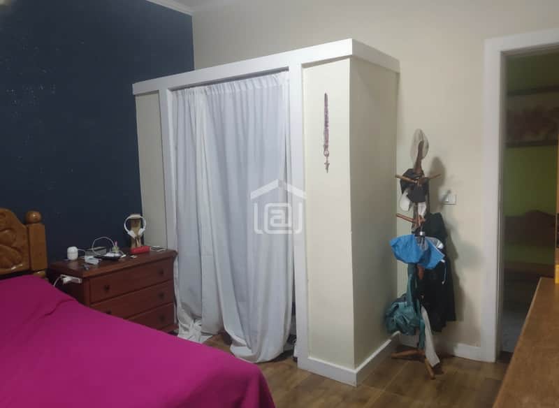 Quarto com cama rosa, cômoda de madeira e guarda-roupa com cortina branca. Cabideiro de madeira ao lado.