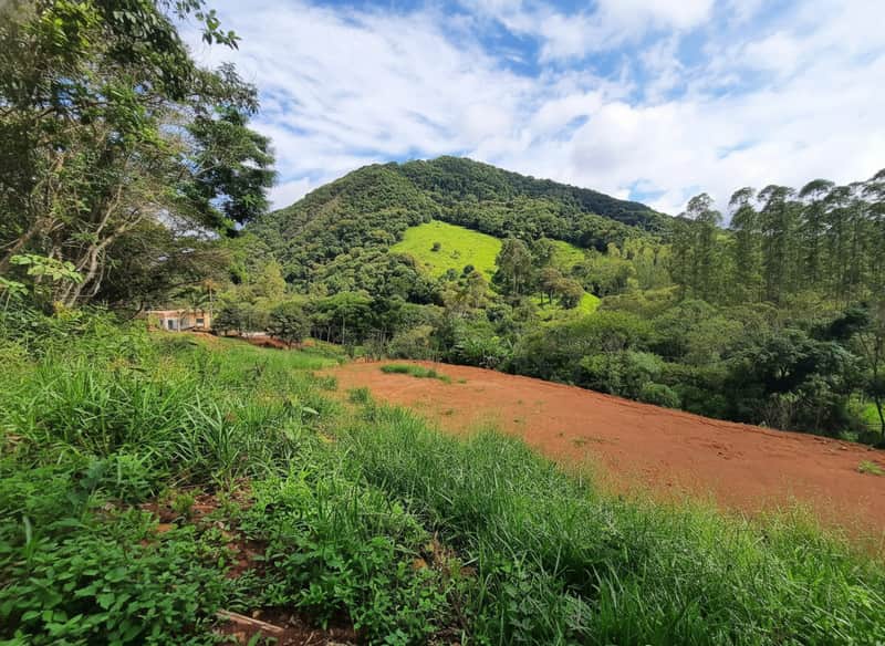 Área externa com morro verde ao fundo. Terreno de terra batida e vegetação rasteira. Casa discreta à esquerda.