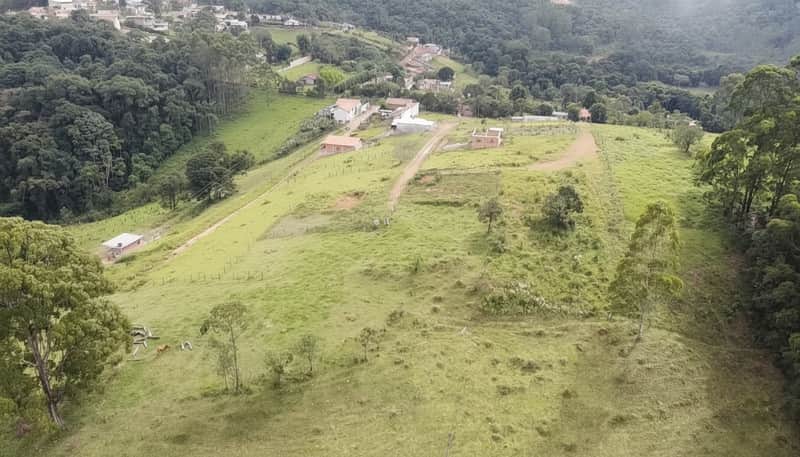 Área externa rural com casas, pasto verde e mata. Vista ampla e campestre, com construções rústicas.