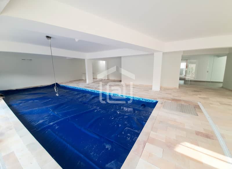 Piscina coberta com capa azul. Ambiente amplo, claro, com piso bege e colunas brancas.
