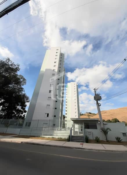Fachada de prédio residencial alto, com portaria, cerca e rua. Céu azul com nuvens ao fundo.