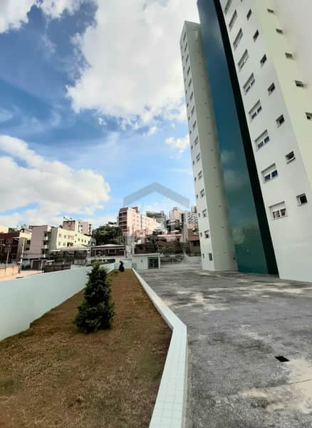 Área externa com gramado e prédio alto com fachada moderna e janelas. Céu azul com nuvens.