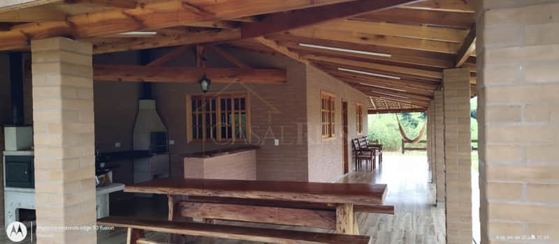 Varanda rústica com churrasqueira, mesa de madeira maciça e colunas de tijolo à vista. Ambiente campestre e aconchegante.