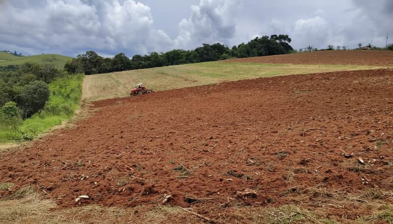 Terra arada em área rural. Trator vermelho ao fundo, preparando o solo para plantio. Céu nublado.