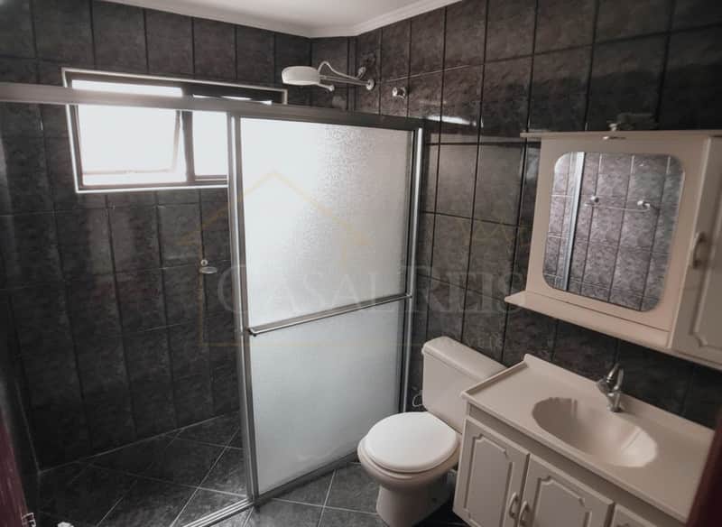 Banheiro com box, pia com armário bege e vaso sanitário. Revestimento cerâmico escuro nas paredes e piso.