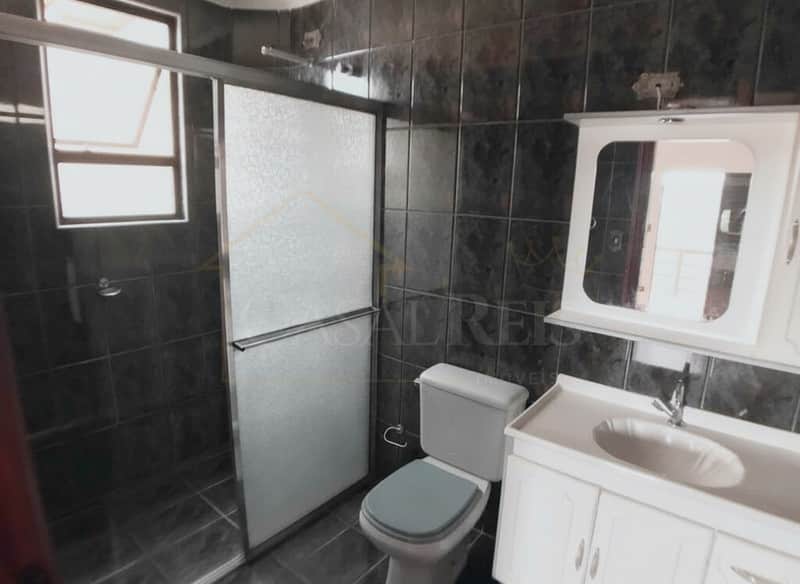 Banheiro com azulejos escuros, box com porta de correr, vaso sanitário e pia com armário branco.