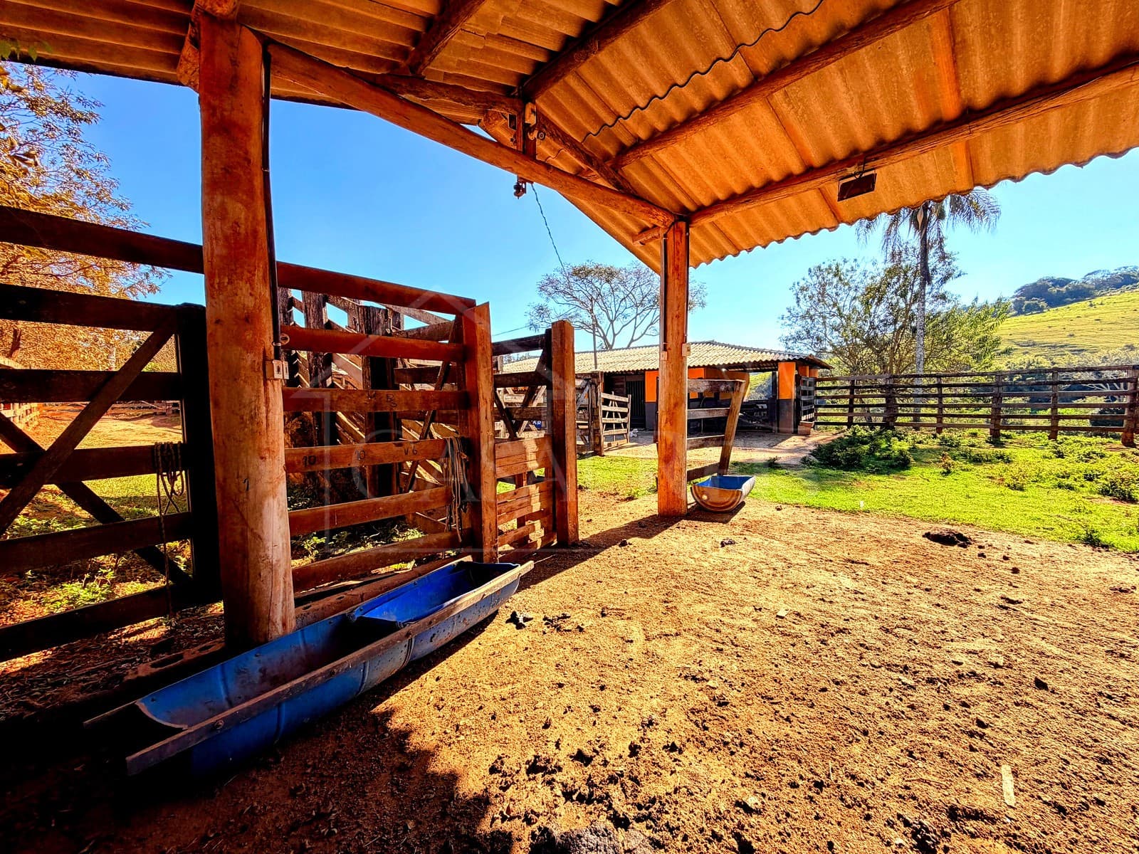 Área externa de fazenda com curral de madeira, cocho azul e telhado rústico sob céu azul.