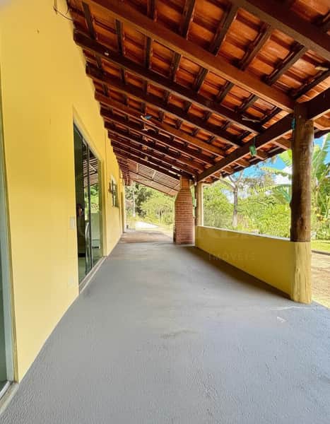 Varanda ampla com piso cinza, pilares de madeira e telhado rústico. Área externa iluminada e arejada.