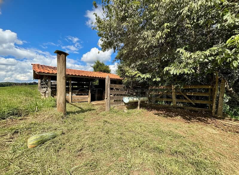 Área externa rural com rancho rústico, cerca de madeira e vegetação. Céu azul com nuvens.