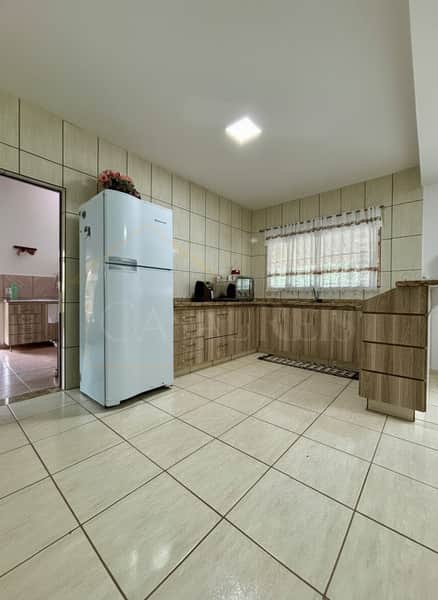 Cozinha ampla com piso cerâmico, armários em madeira clara e geladeira branca. Janela com cortina ilumina o ambiente.