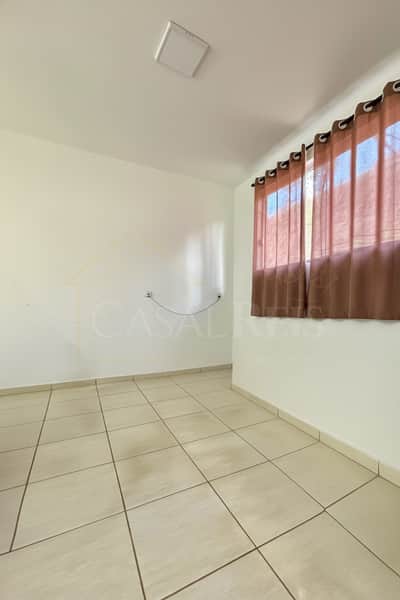 Quarto claro com piso cerâmico, cortina marrom na janela e paredes brancas. Ambiente arejado e iluminado.