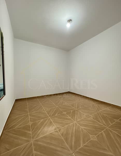 Quarto vazio com piso cerâmico marrom e paredes brancas. Iluminação central.