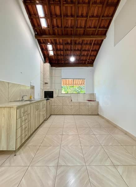 Cozinha ampla com armários claros, churrasqueira e piso cerâmico. Teto rústico com vigas de madeira e iluminação central.